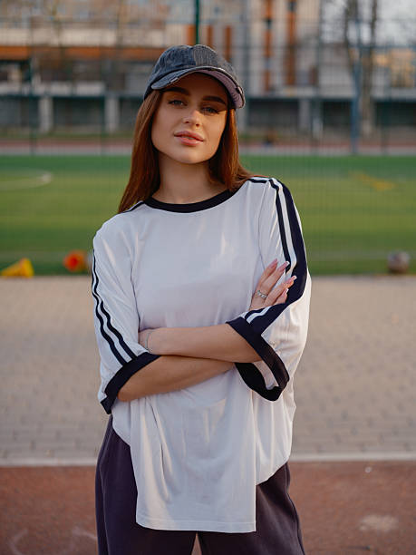 Female model wearing a white t-shirt and baseball cap standing on an athletic court. Urban background with residential buildings and green turf during sunset.