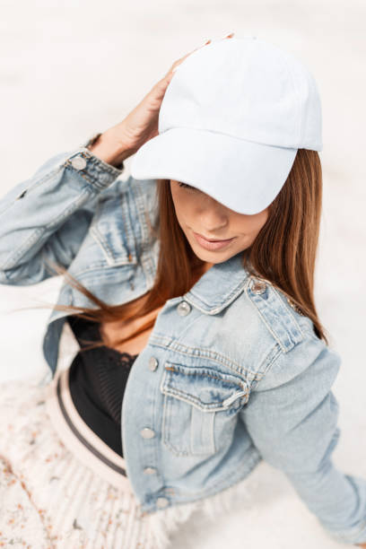 Stylish pretty young woman in a fashionable denim jacket in a blue cap sits on the white sand and rests on summer holidays